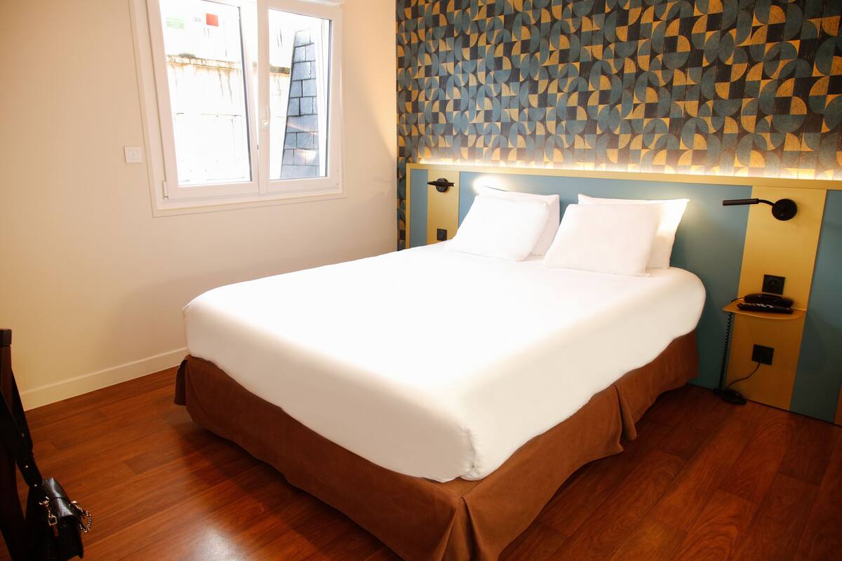 A neatly made bed with white linens, a wooden floor, and a patterned wall behind it.