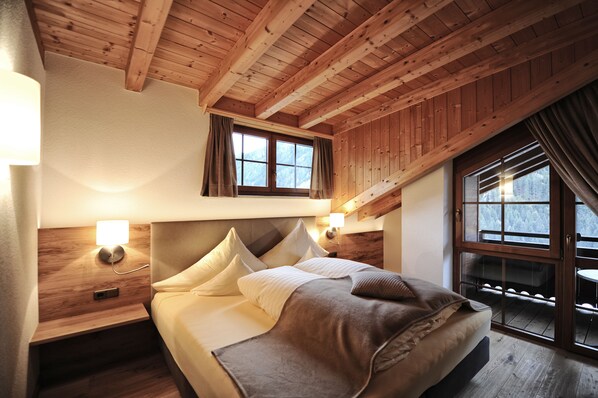 Ein Schlafzimmer mit Holzboden, einem Bett mit Kissen, einem Nachttisch mit Lampe und einem Fenster mit Vorhängen.