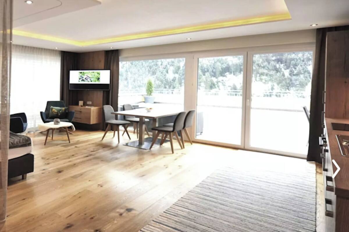Una sala de estar moderna con piso de madera, un área de comedor con mesa y sillas, un sofá, un televisor y un ventanal con vista a árboles.