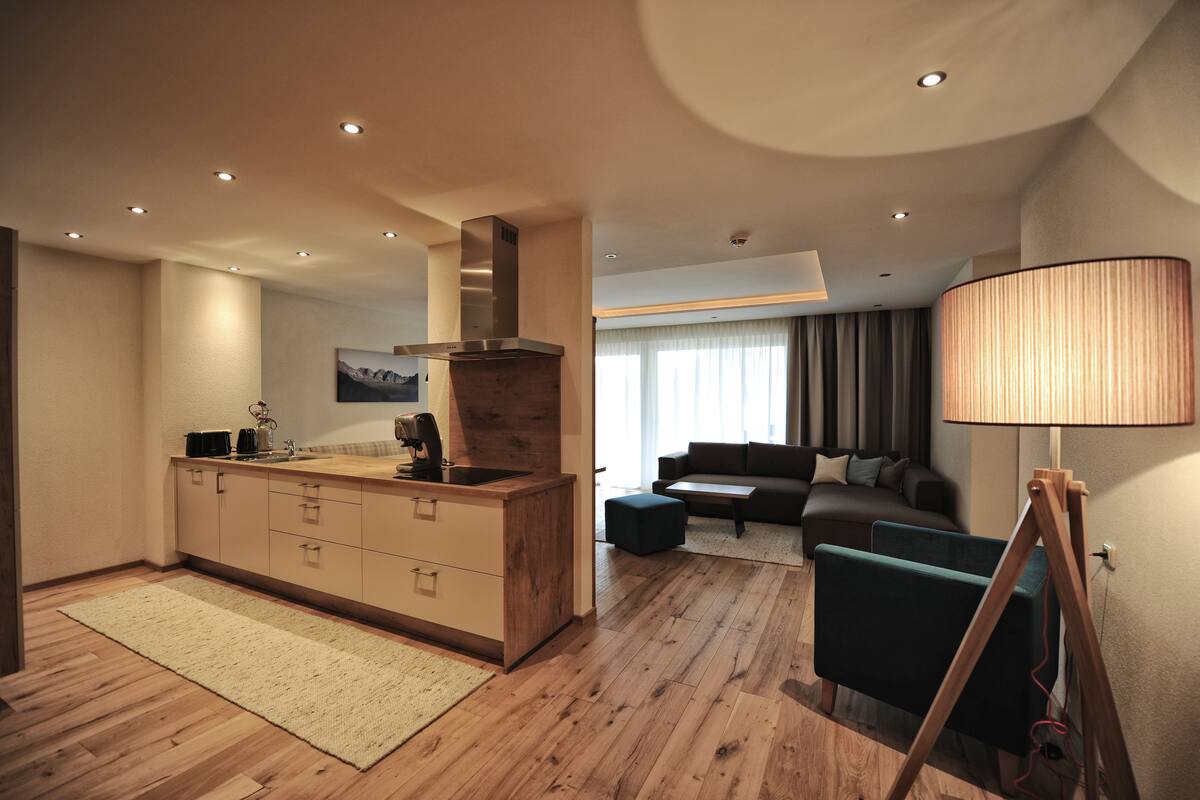 Una cocina moderna con armarios de madera y una estufa integrada, un espacio de sala de estar con un sofá y una mesa de centro, y una lámpara sobre un trípode.