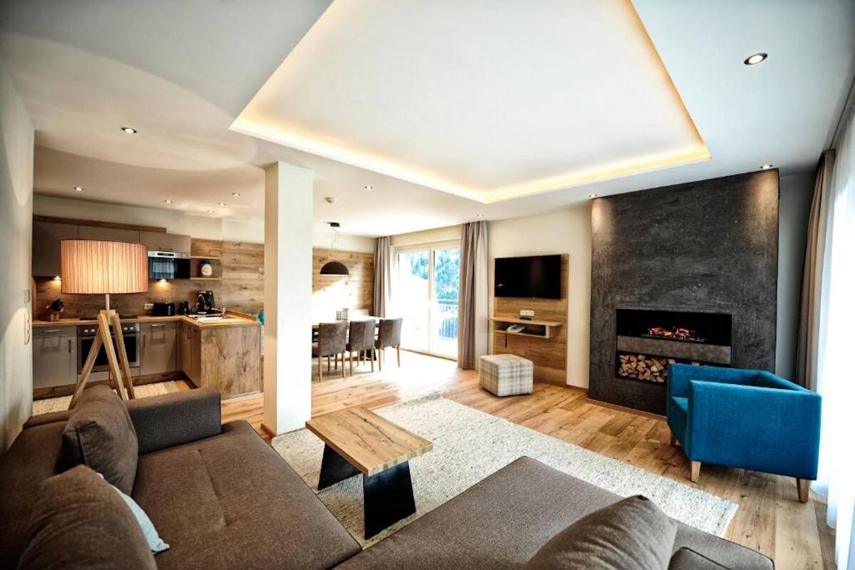 Una sala moderna con un sofá, un televisor y una chimenea. Se observa una cocina al fondo con zona de comedor.