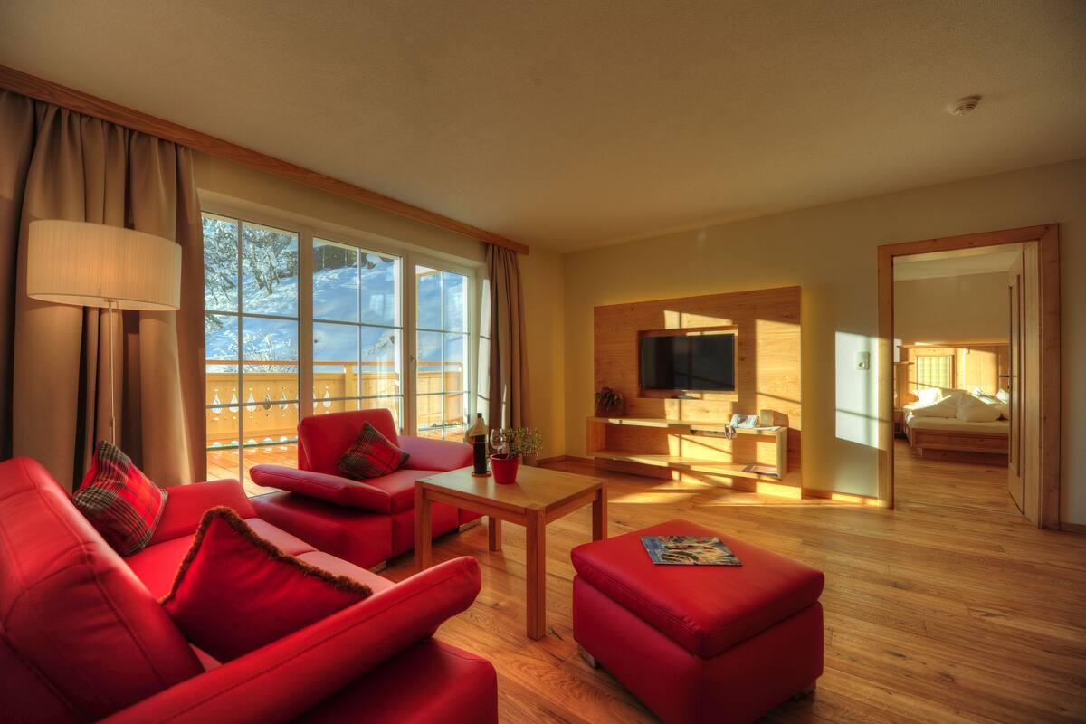 Una sala de estar moderna con sofás rojos, una mesa de centro de madera y un televisor. Amplias ventanas ofrecen vistas al exterior.