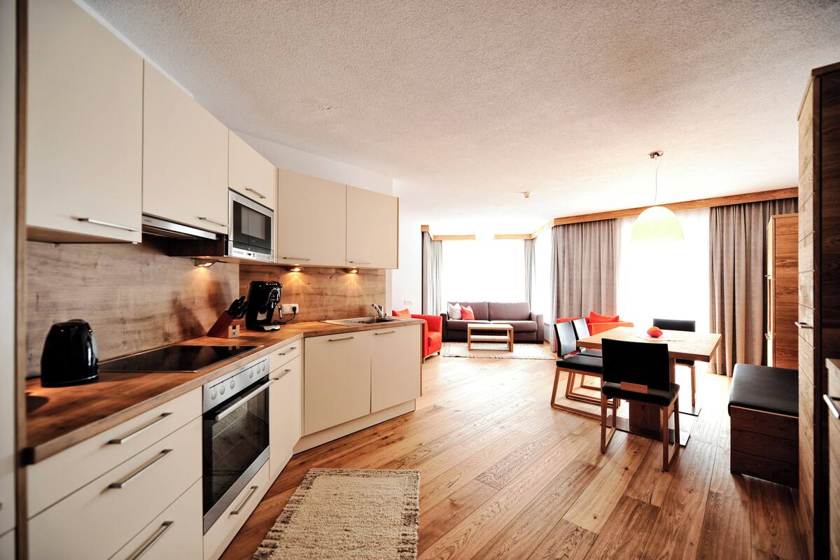 Cocina moderna con gabinetes blancos, horno integrado y microondas. Hay un comedor con mesa y sillas de madera, y un sofá con cojines.