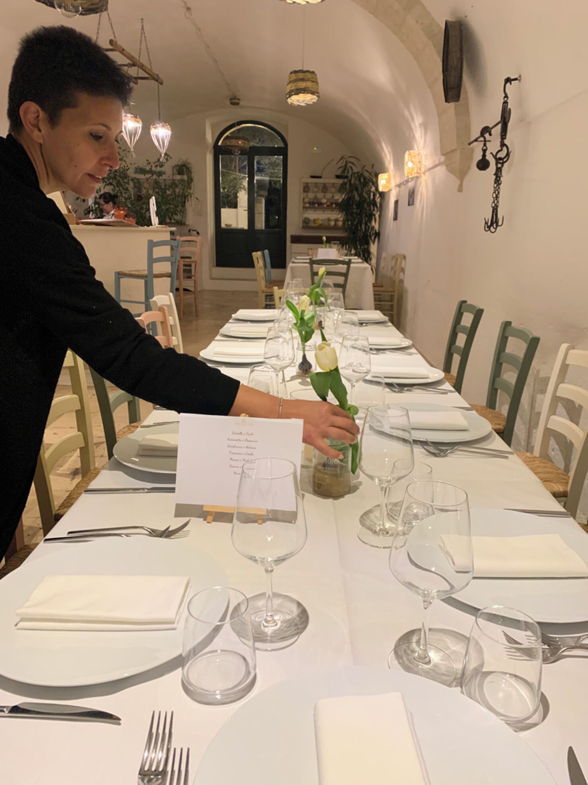 Mesa de comedor dispuesta con manteles blancos, platos y copas de vino, con una persona arreglando flores.