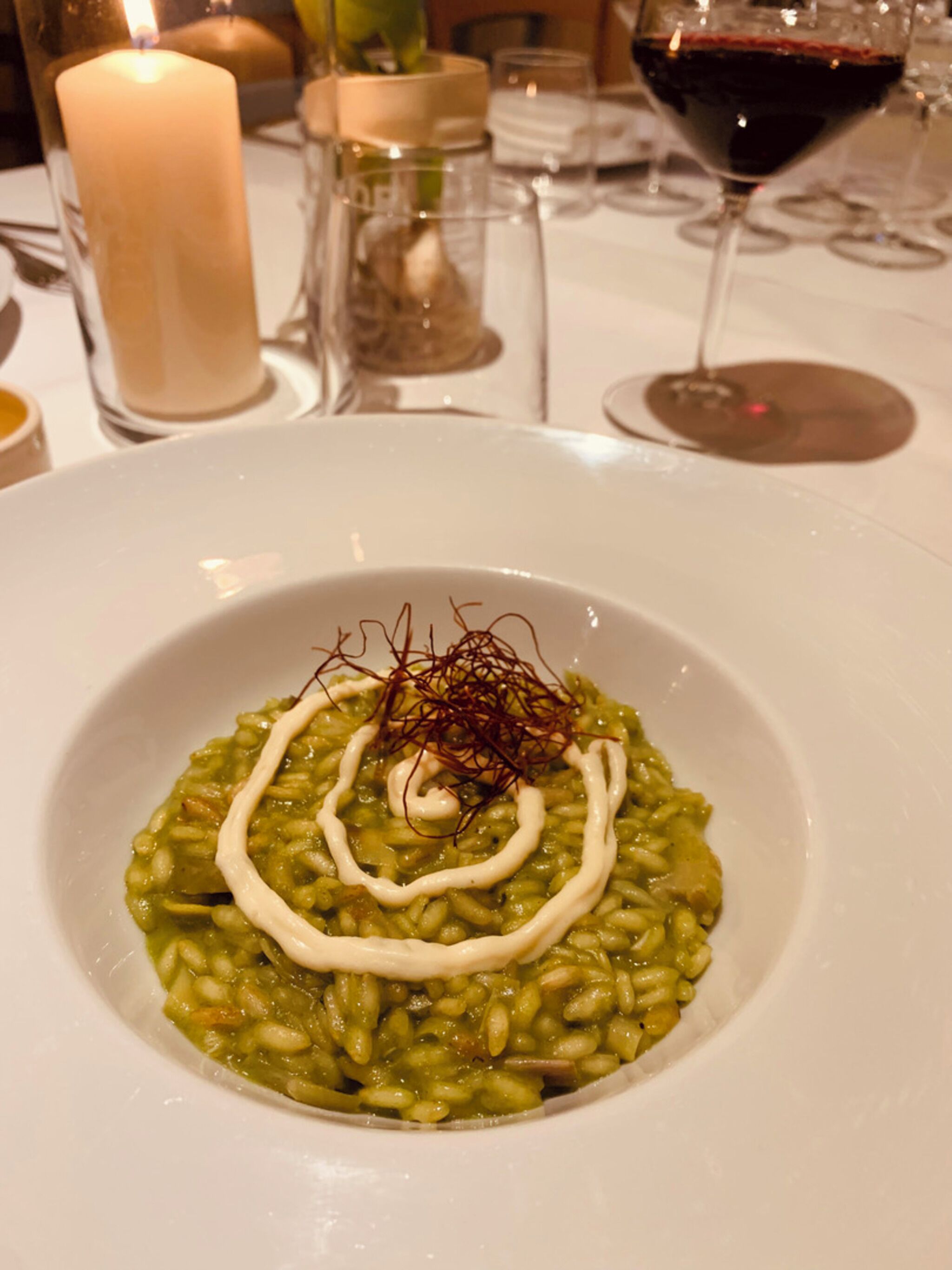 En tallrik med grön risotto, vit sås och saffransdekoration, placerad på ett bord med ljus och vinglas.