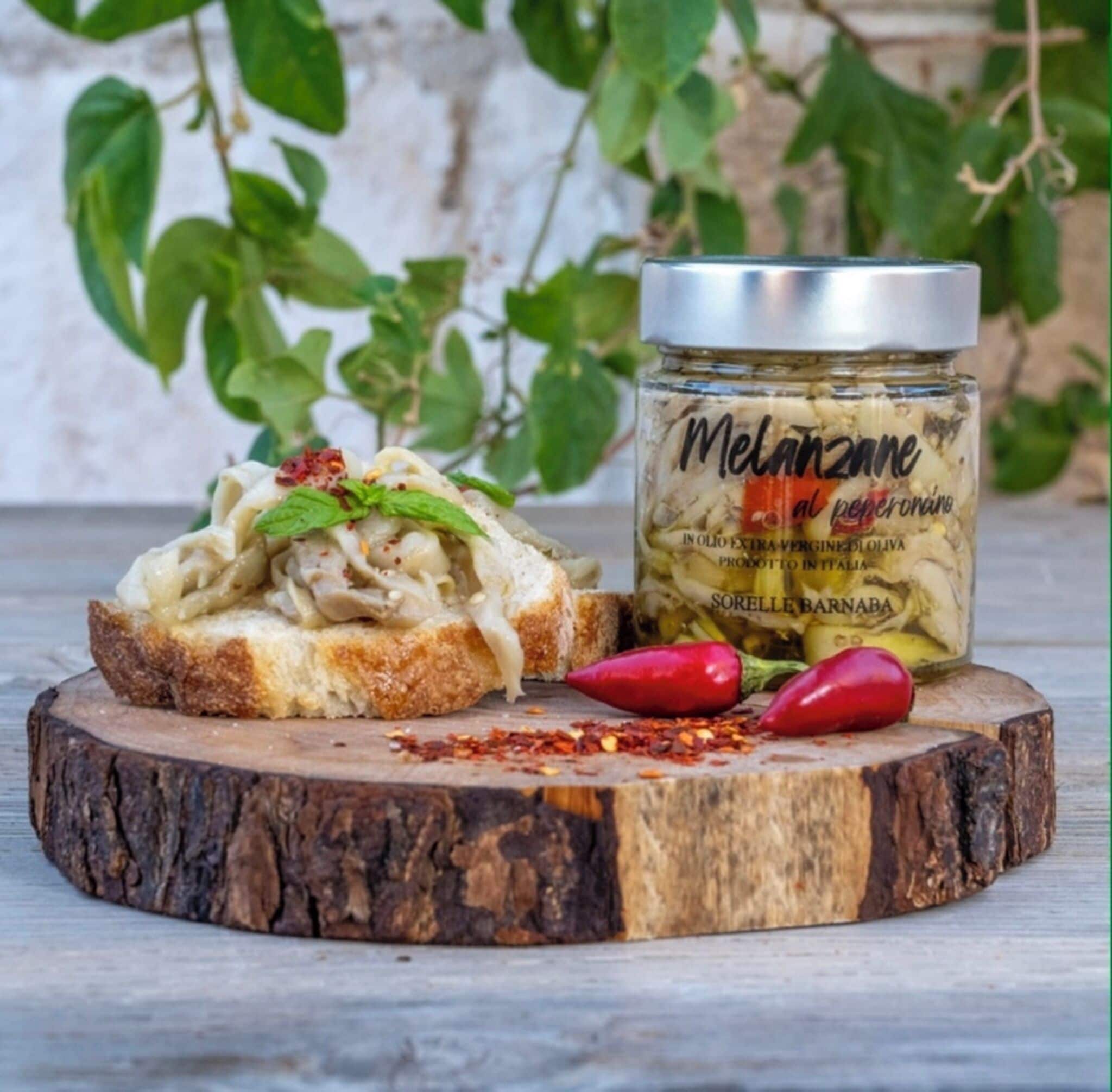 Ein Glas Melanzani al peperonata auf einer Holzbrett mit einer Brotscheibe, belegt mit Soße und Chiliflocken.