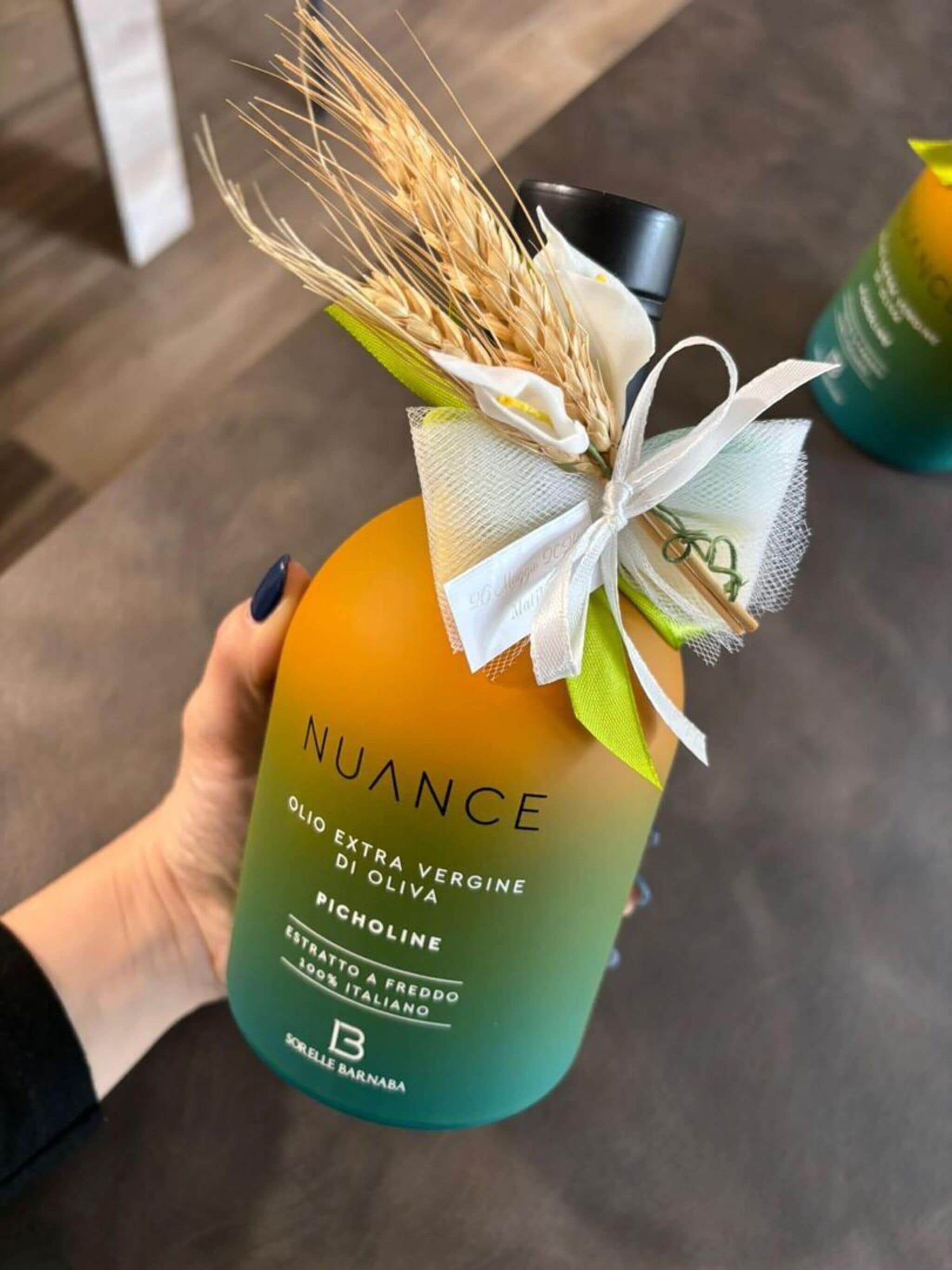 Una mano sostiene una botella de aceite de oliva Nuance con etiqueta degradada, una cinta blanca y espigas de trigo secas.