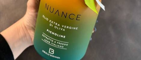 Una mano sostiene una botella de aceite de oliva Nuance con etiqueta degradada, una cinta blanca y espigas de trigo secas.