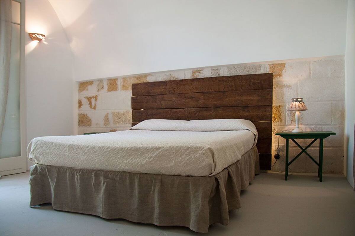 Une chambre à coucher avec une tête de lit en bois, un lit recouvert d’une couverture beige et une petite table avec une lampe.