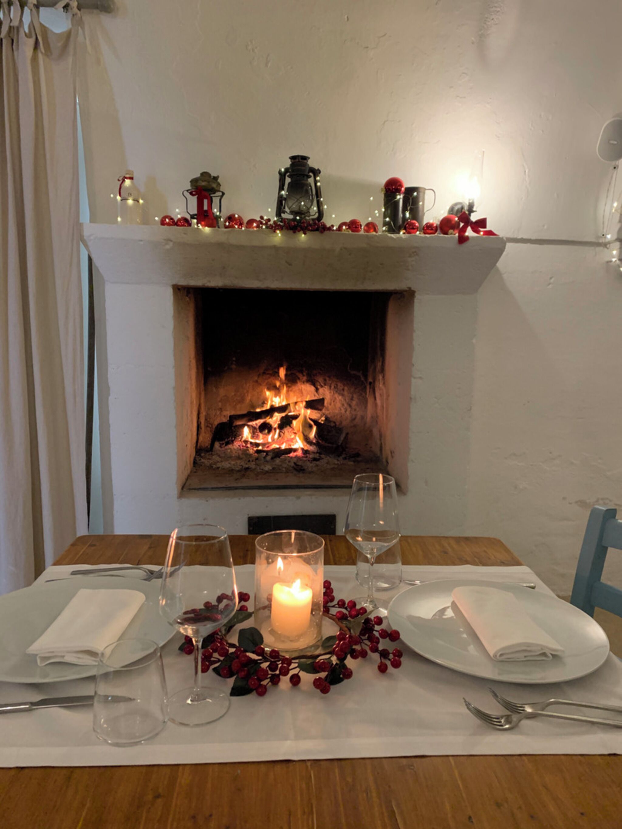 A dining table set for two with a lit candle centerpiece, a fireplace in the background, and festive decorations.