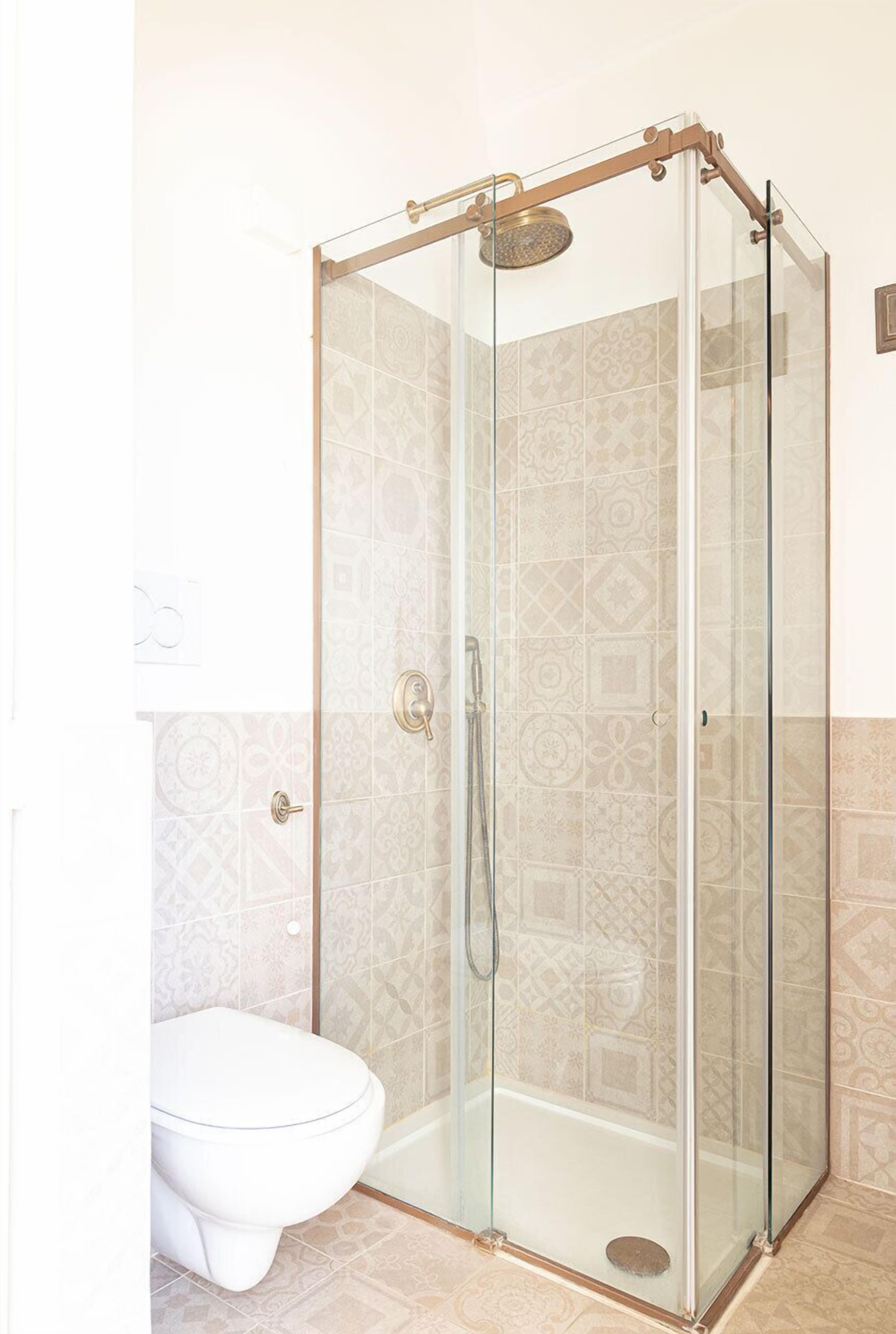 Ducha con mampara de vidrio, cabezal de ducha y retrete en un baño con azulejos estampados.