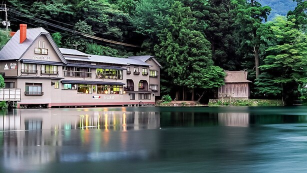 A large, multi-story building with a balcony, situated by a calm lake, surrounded by dense trees.