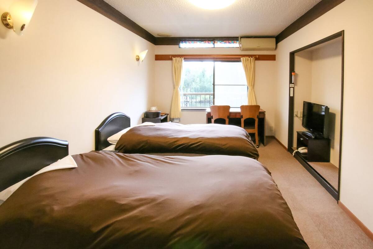 Una habitación de hotel con dos camas, un escritorio con sillas, un televisor y una ventana con cortinas.