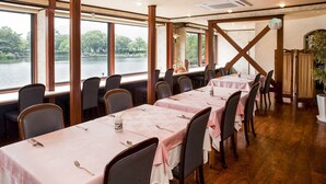 A restaurant with multiple tables, each with a pink tablecloth and set with cutlery. Large windows offer a view of a lake and trees.