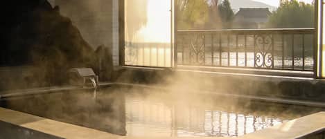 A hot spring bath with steam rising, a stone wall, and a view of a traditional building through a window.