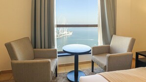 Quarto de hotel com duas poltronas, uma mesa redonda pequena e uma cama. Uma grande janela oferece vista para um corpo de água e uma roda gigante ao longe.