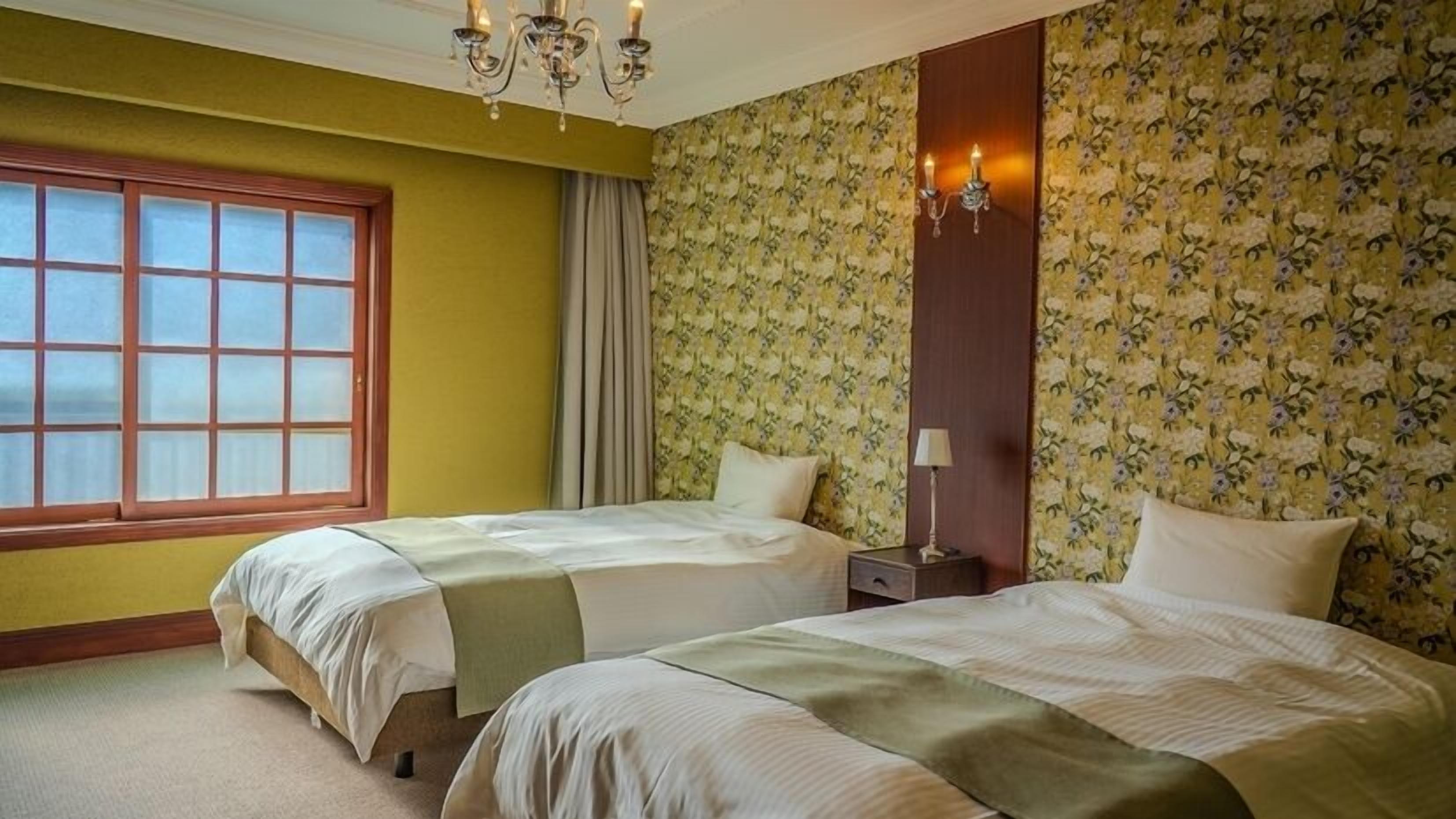 A hotel room with two beds, a window, a chandelier, and floral wallpaper.
