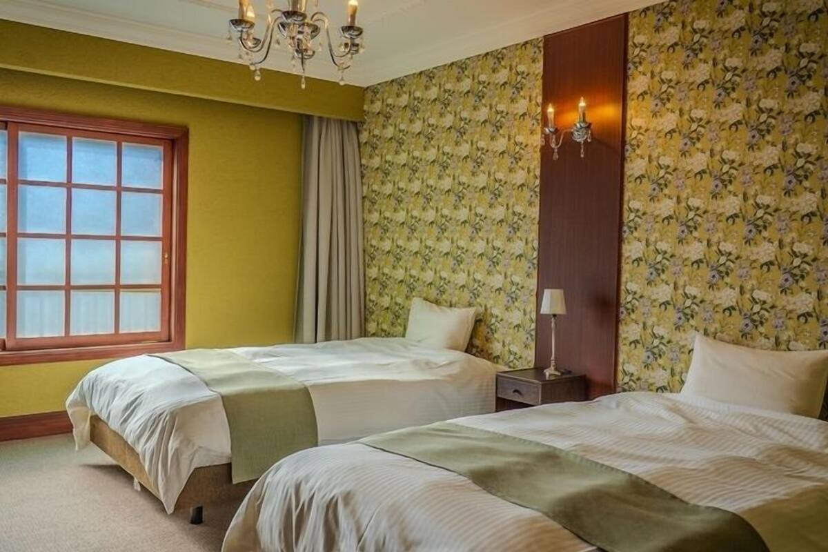 A hotel room with two beds, a window, a chandelier, and floral wallpaper.