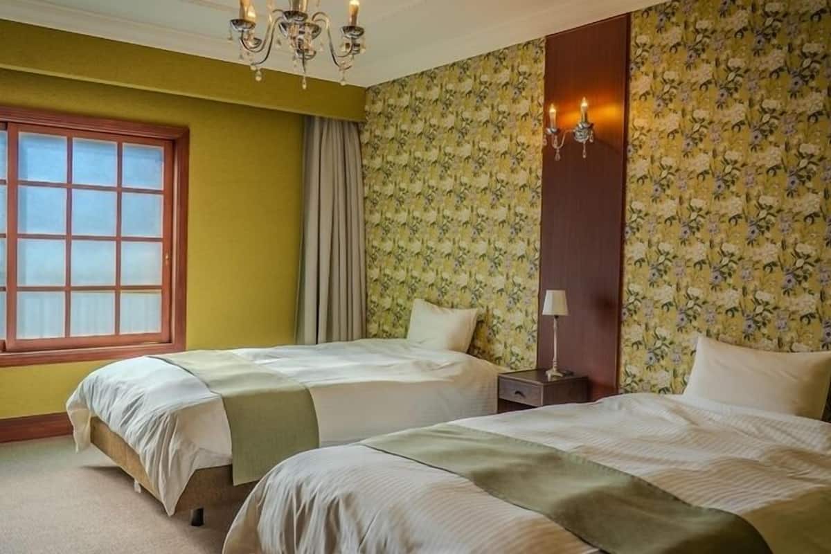 A hotel room with two beds, a window, a chandelier, and floral wallpaper.