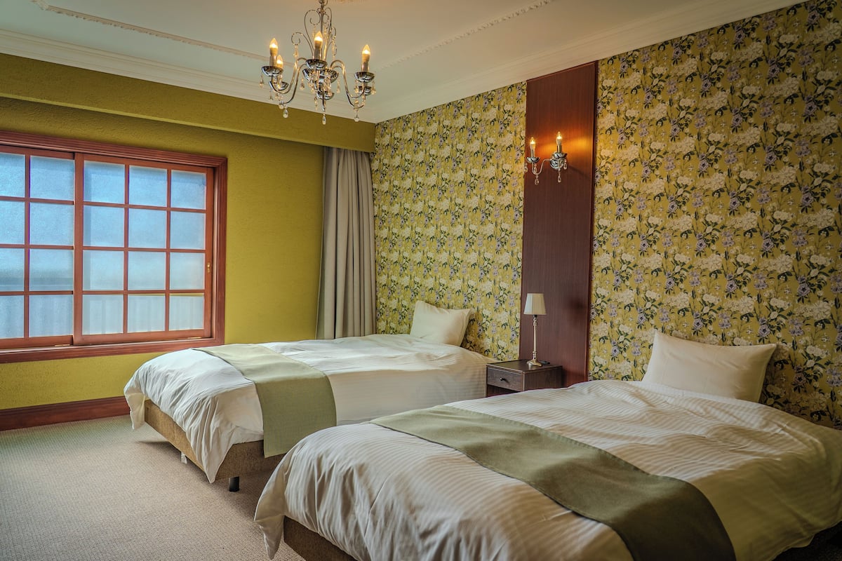 A hotel room with two beds, a large window, floral wallpaper, and a chandelier.