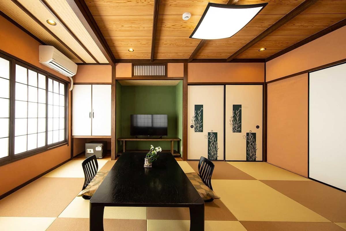 A traditional Japanese-style room with tatami flooring, a tatami mat table, a television, sliding doors, and a ceiling light fixture.