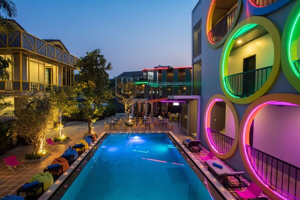 A swimming pool with lounge chairs and tables, surrounded by buildings with colorful circular windows. - White Rabbit Hostel - Adults Only (Siem Reap)