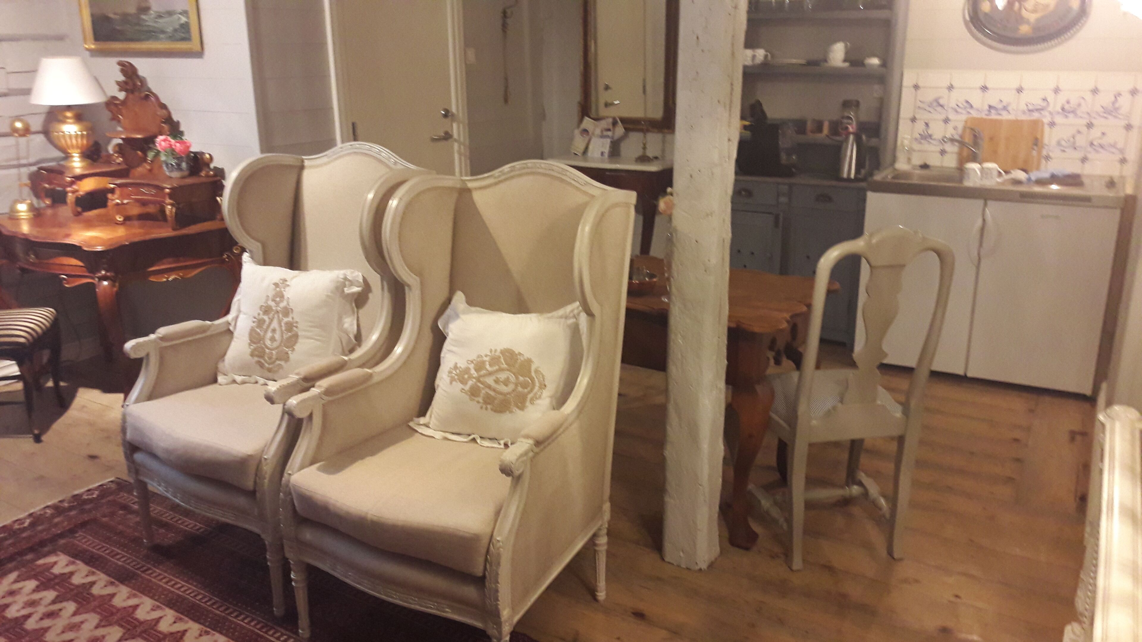 Una habitación con un sillón tradicional, una mesita pequeña y una zona de cocina al fondo.