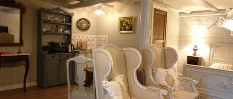 A traditional room with a wooden floor, a white bed with a decorative headboard, and a white armchair with a cushion. There is a wooden cabinet with glassware and a clock on the wall.