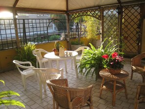 Un patio techado con muebles de mimbre, plantas en macetas y una mesa con una jarrón y vasos.