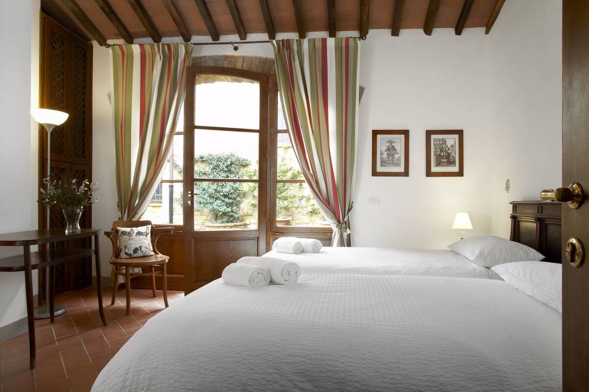 Habitación de hotel con dos camas, un escritorio, una silla, una ventana con cortinas y dos cuadros en la pared.