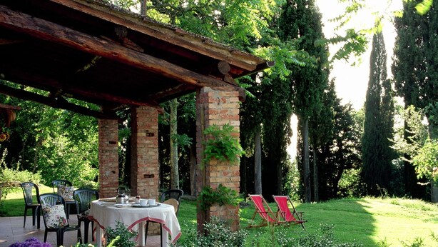 Un área de estar al aire libre, techada, con una mesa y sillas, rodeada de abundante vegetación y árboles.