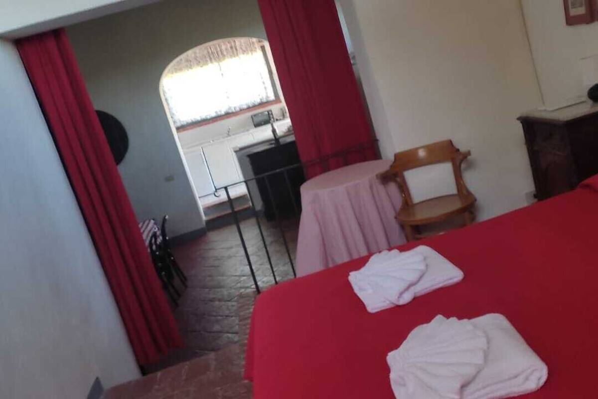 Un dormitorio con una colcha roja, dos toallas dobladas, una silla de madera, una mesita y un ventanal con cortinas.