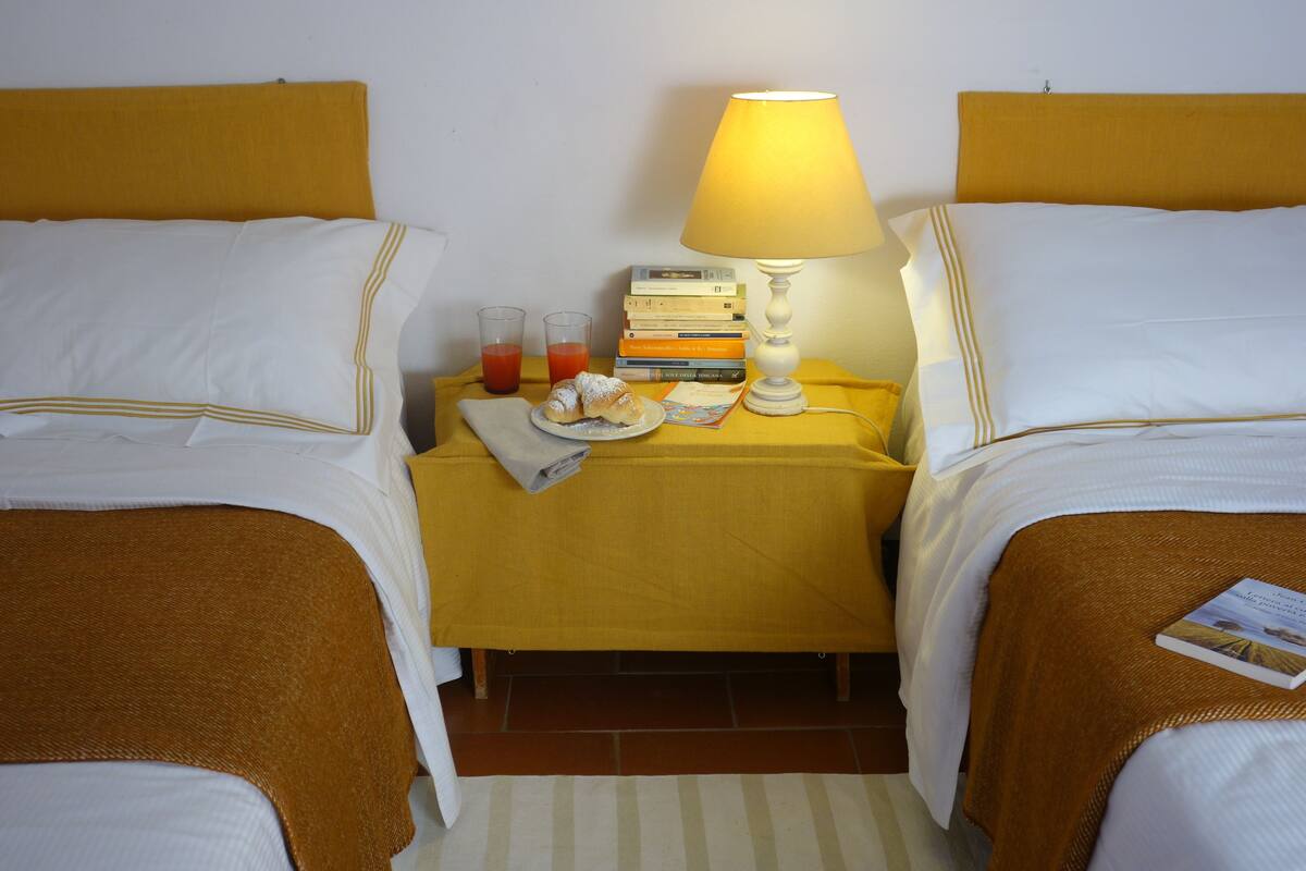 Habitación de hotel con dos camas, una mesita de noche con lámpara, libros, un plato con comida y vasos de jugo.