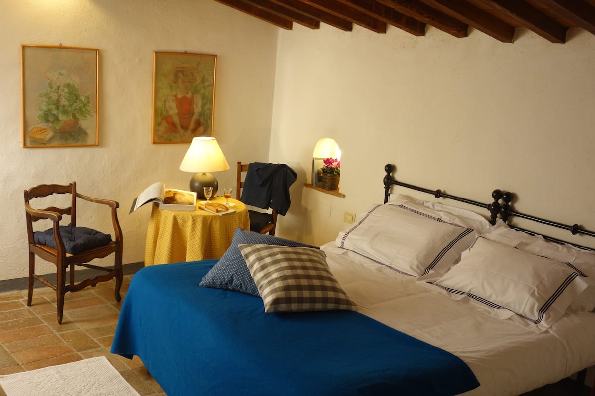 Un dormitorio con una cama, una silla, una mesa con lámpara y dos cuadros en la pared.