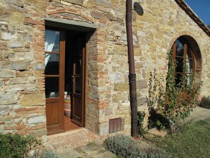 Un edificio de piedra con puerta y ventanas de madera, un pequeño jardín con arbustos y un sendero de ladrillo que lleva a la entrada.