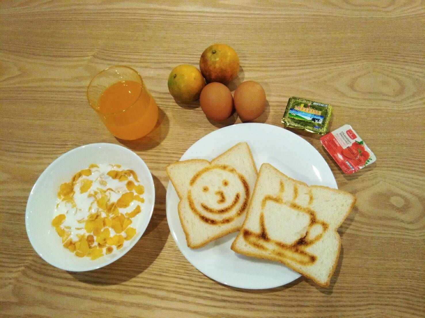 A wooden table with breakfast items including toast with smiley faces, a bowl of cereal, a glass of orange juice, eggs, and a packet of strawberry jam.