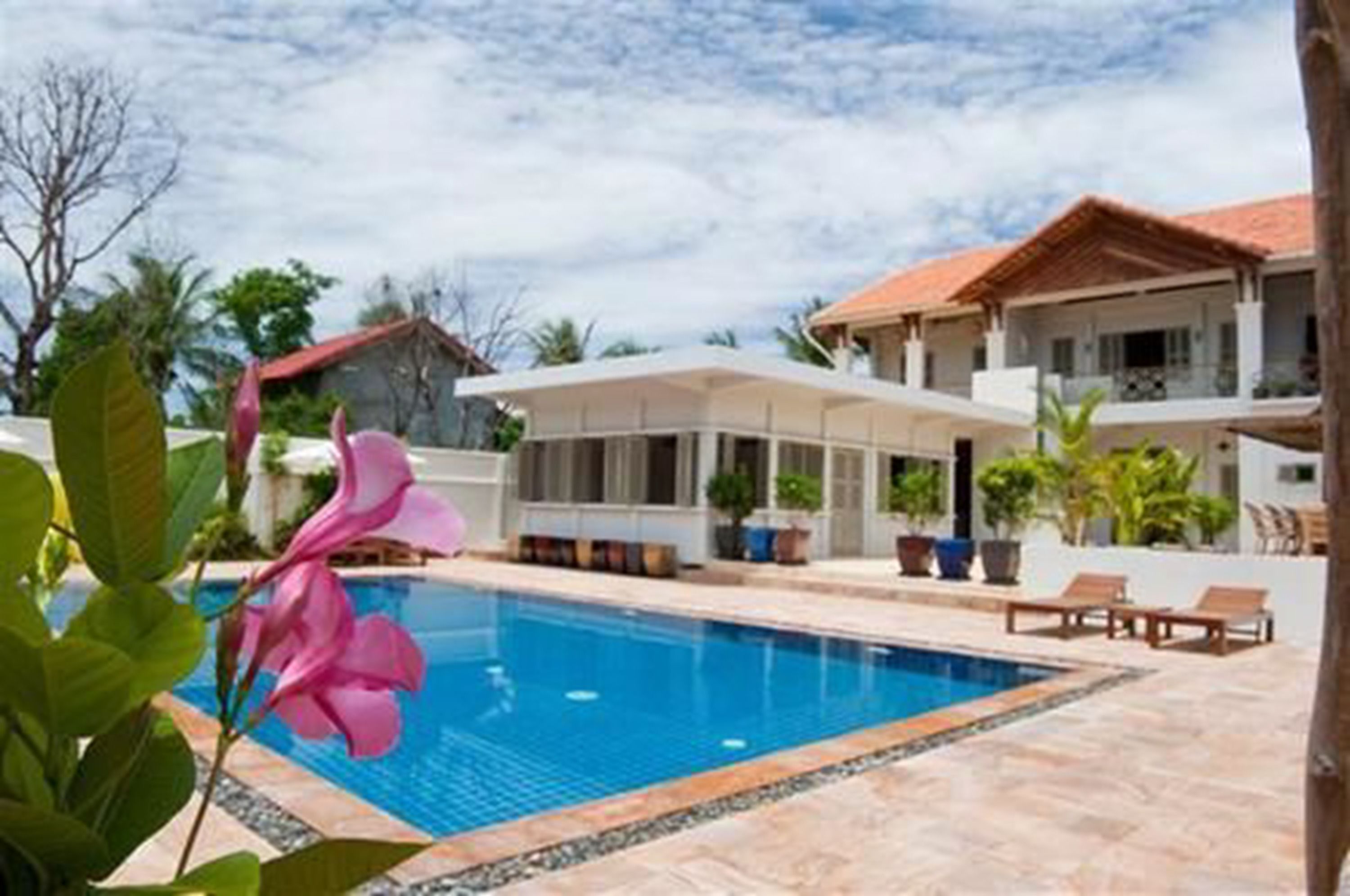 A swimming pool with a tiled deck, a covered seating area, and a two-story building with a red-tiled roof.