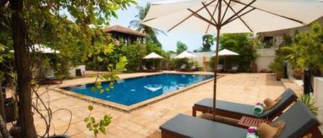 A swimming pool with lounge chairs and umbrellas, surrounded by greenery and a tiled patio.