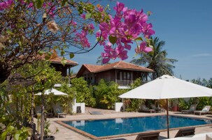 Un área de piscina con sillas de descanso y sombrillas, rodeada de exuberante vegetación y una casa con tejado de baldosas.