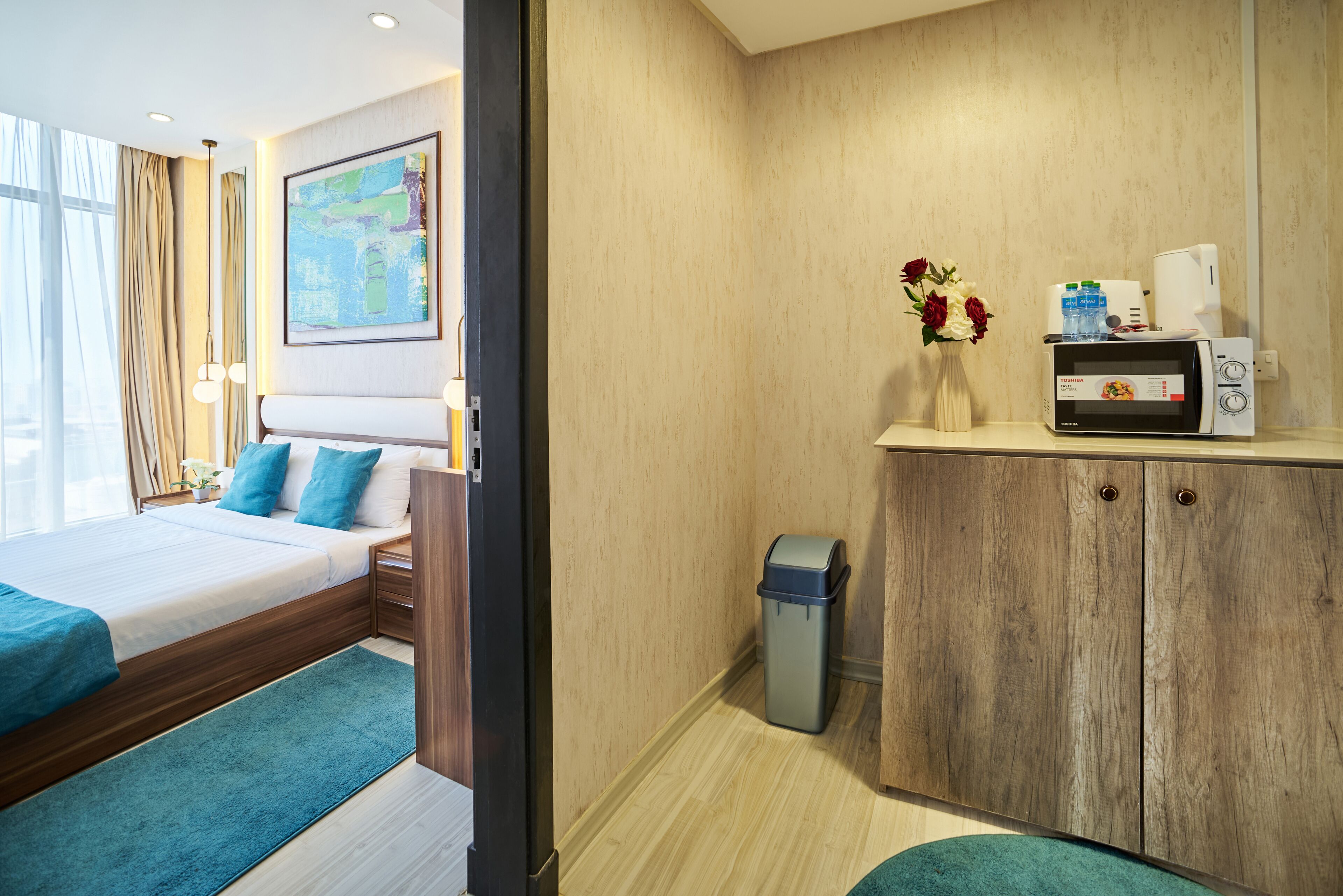 A hotel room with a bed, a small desk with a microwave, a trash bin, and a vase with flowers.