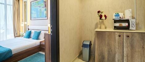 A hotel room with a bed, a small desk with a microwave, a trash bin, and a vase with flowers.