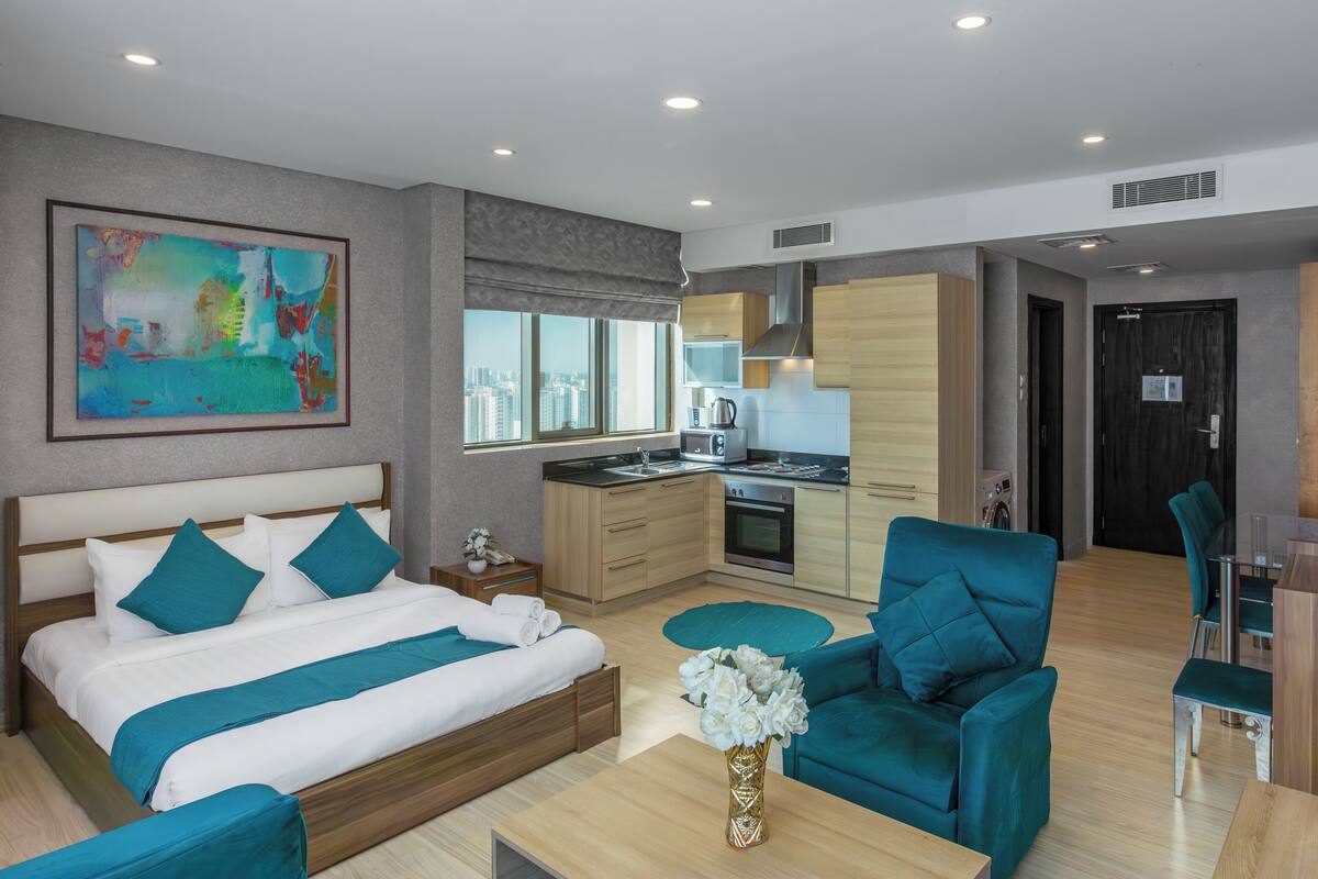 A modern hotel room with a bed, a sitting area, a kitchenette, and a view of the city.