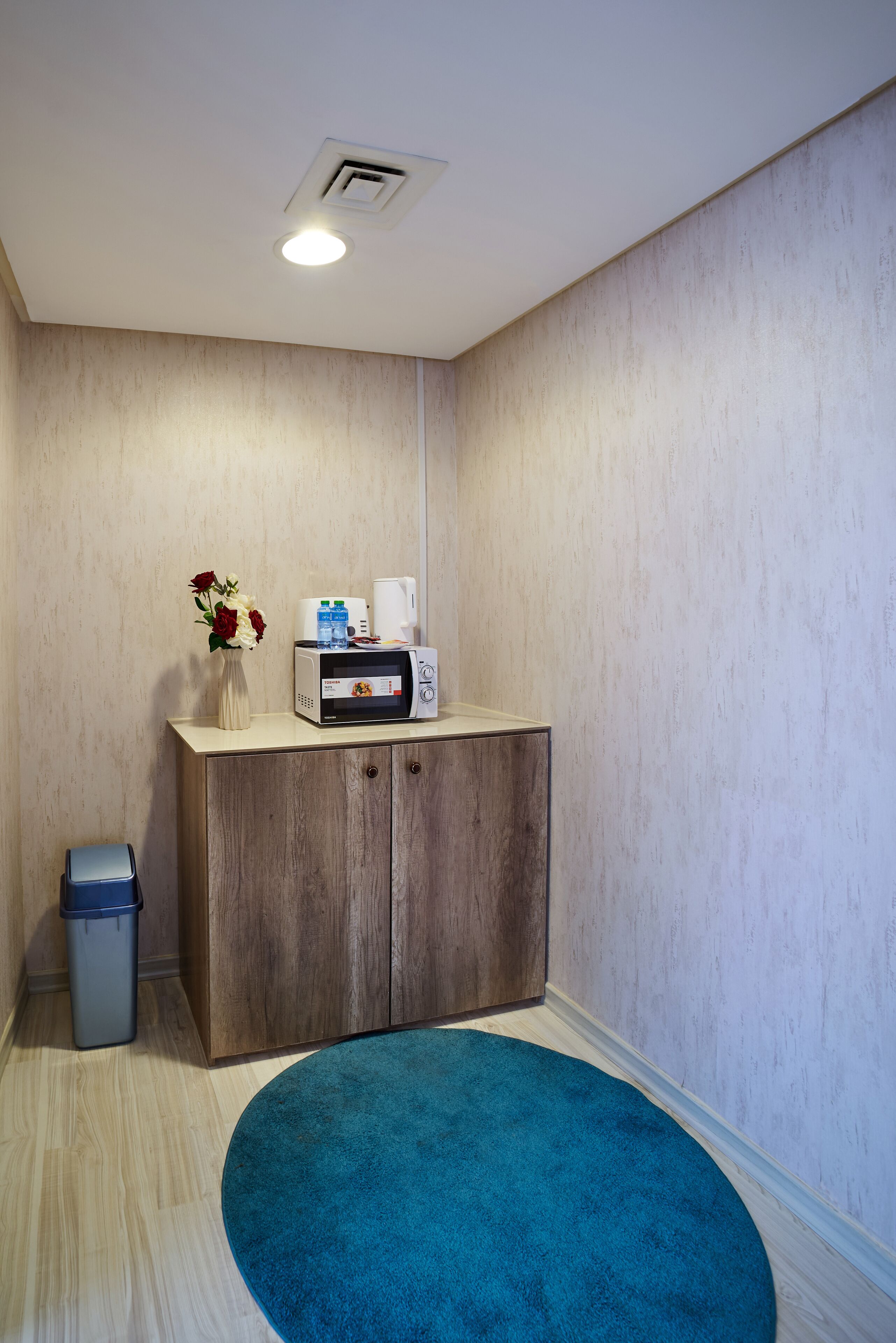 A small, private room with a wooden cabinet, a microwave, a vase with flowers, and a blue rug.