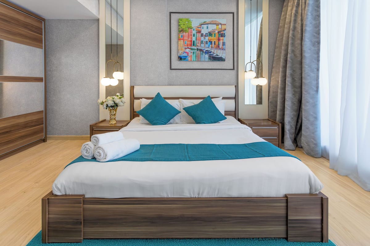 A neatly made bed with white and teal bedding, two teal pillows, and a rolled white towel on a wooden platform bed. There is a small table with a vase of flowers and a lamp beside the bed.