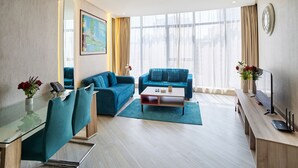 Habitación de hotel moderna con dos sofás azules, una mesa de comedor de vidrio, un escritorio de madera y un amplio ventanal con cortinas.