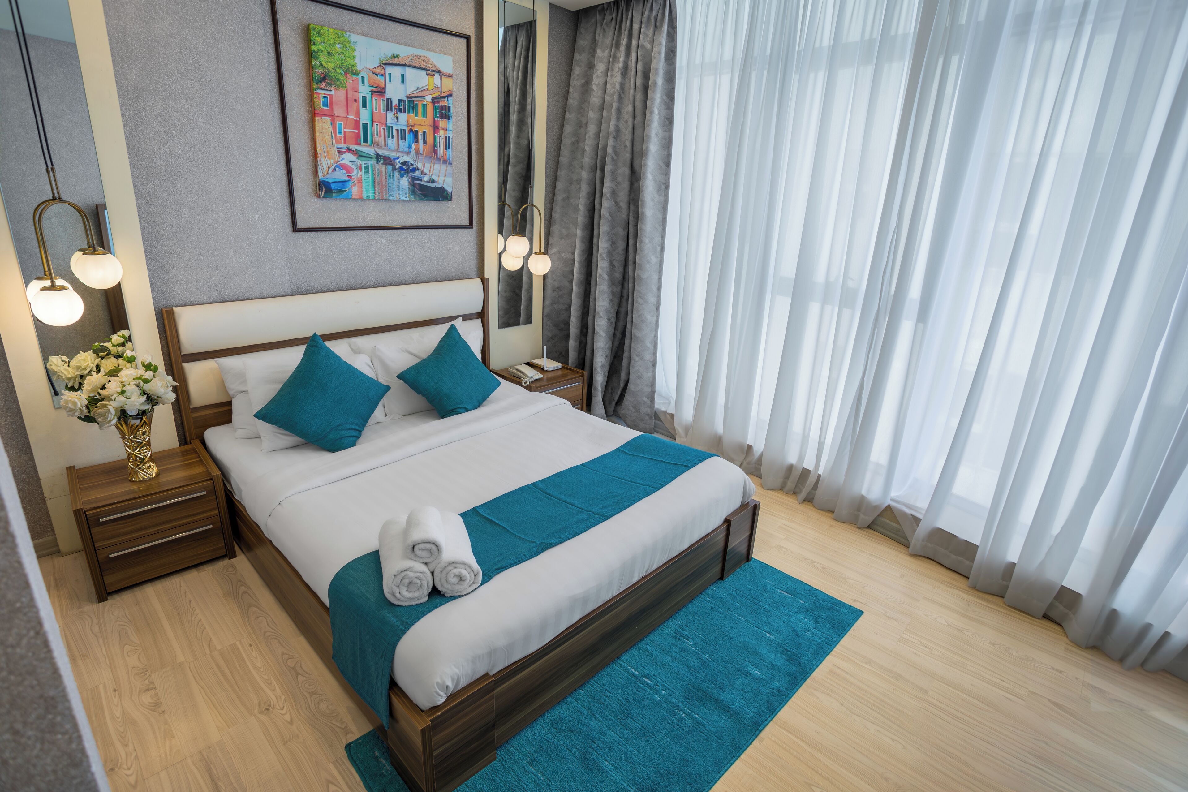 Una cama bien hecha con una colcha verde azulada y almohadas, una mesita de noche de madera con un florero y un cuadro en la pared.