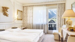 A hotel room with a bed, a desk with a lamp, a chair, and a view of mountains through a window.