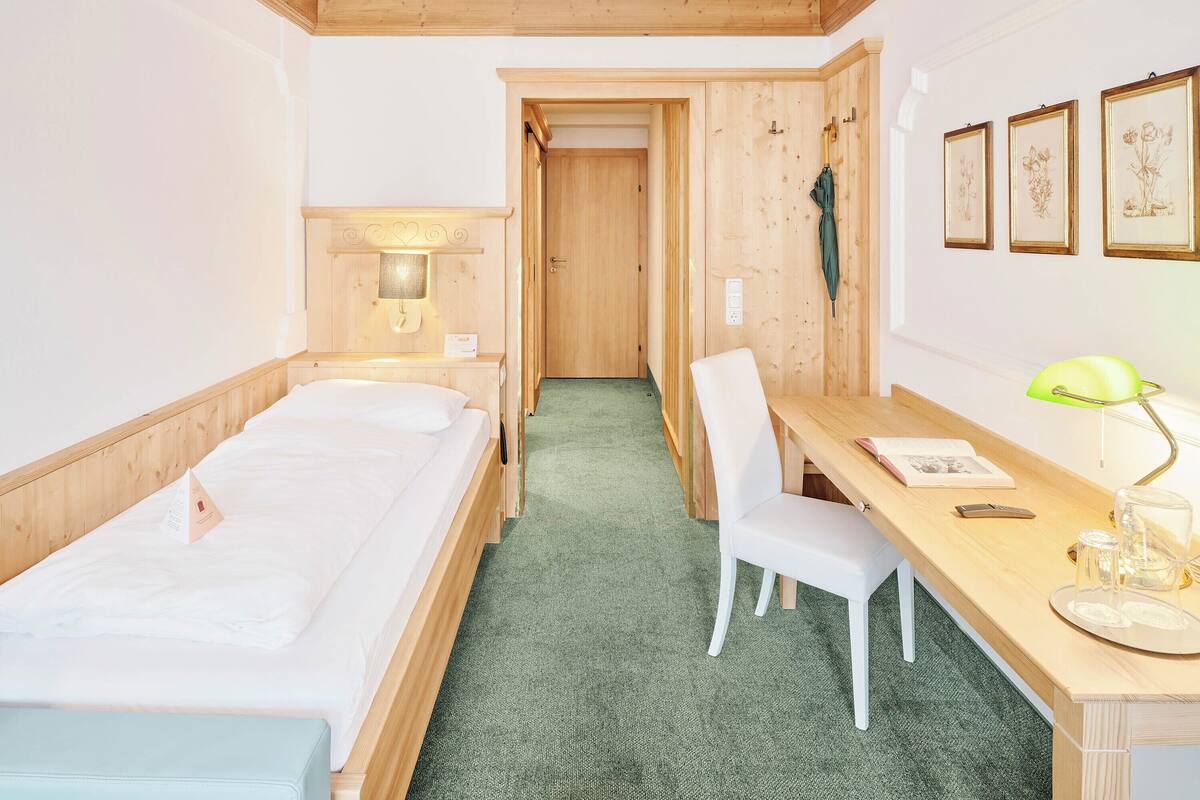 Hotellihuone, jossa on kaksi erillistä sänkyä, työpöytä tuolilla, vihreä matto ja seinällä kehyksiin asetettuja kuvia.