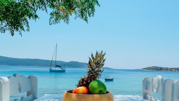A table with a wooden bowl containing fruits and a sailboat in the background. - Klima Paradise (Samos)