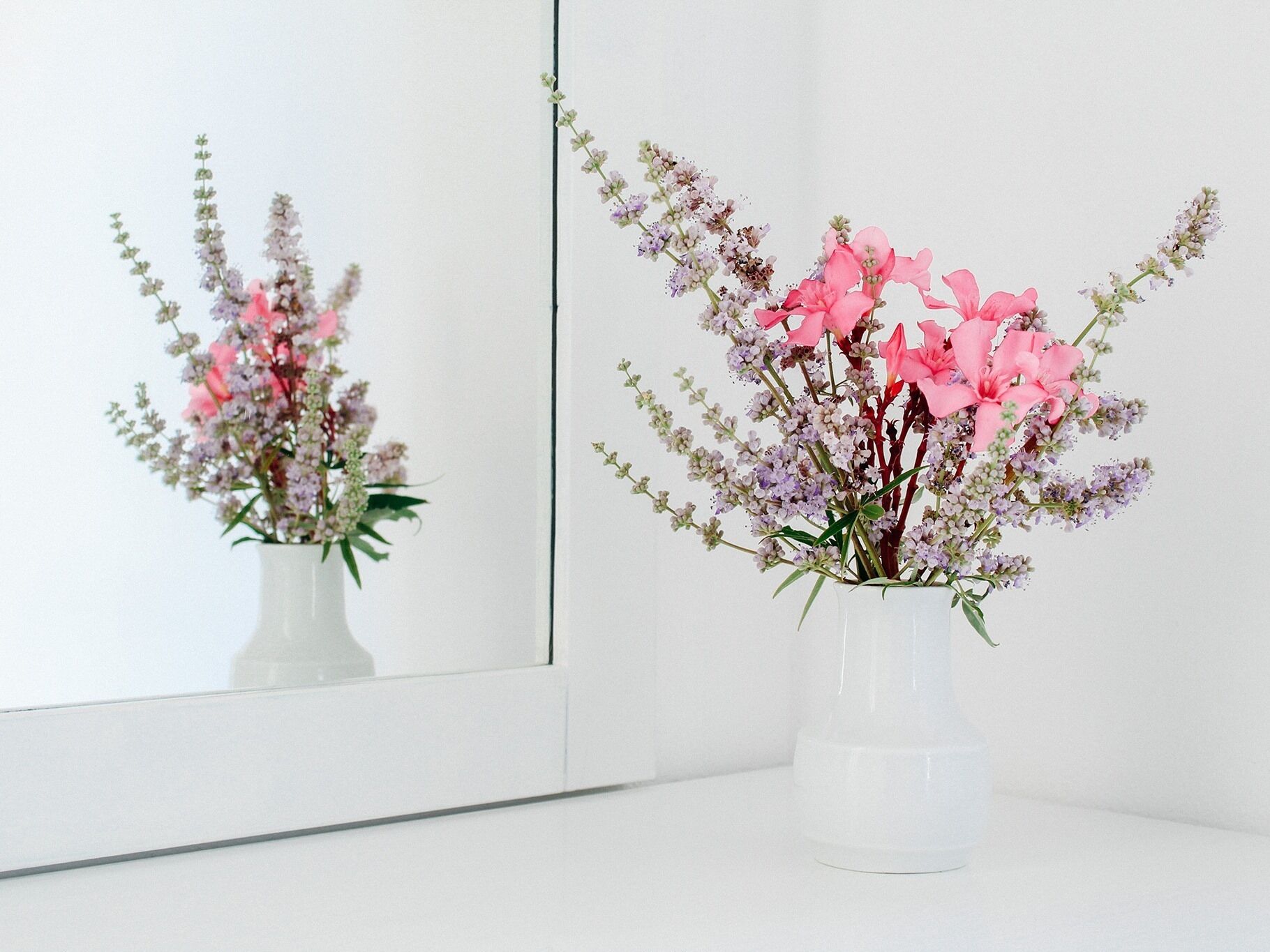 Un florero blanco con flores rosadas y púrpuras sobre una superficie blanca.